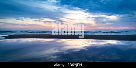 Una tranquilla spiaggia al tramonto, con onde calme che si infrangono sulla riva. Il cielo è dipinto con delicate sfumature di blu, rosa e arancione, che riflettono Foto Stock