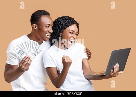 Una coppia nera emozionante che celebra la vittoria con notebook e contanti Foto Stock