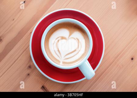 Vista dall'alto della tazza da caffè con latte art a forma di cuore su piattino rosso e tavolo in legno. Vista dall'alto, disposizione piatta. Foto Stock