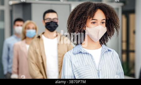 Nuovo controllo normale e della temperatura prima del lavoro. I dipendenti multirazziali millenari nelle maschere protettive sono in linea con il distanziamento sociale in ufficio Foto Stock