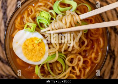 Primo piano di ramen piccante in vivace brodo rosso-arancio, con anelli di porro, uova sode tagliate a metà e bacchette di legno che sollevano una porzione di tagliatelle. Foto Stock