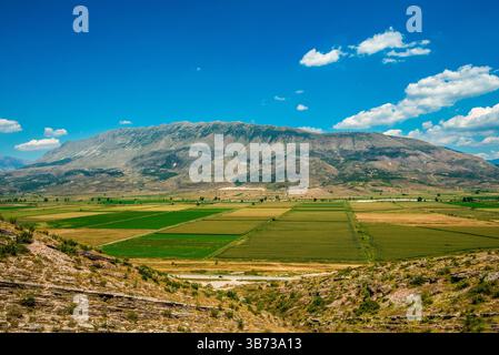 Spazia panoramica delle creste calcaree di Nemercka che salgono sopra i terreni agricoli di patchwork e una strada tortuosa a valle sotto un cielo albanese abbellito da nuvole Foto Stock