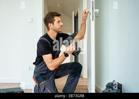 Uomo che installa porte interne con trapano elettrico. Lavoratore professionista che indossa l'uniforme da lavoro con maniglia bianca per porta con cacciavite a batteria in apartmen Foto Stock