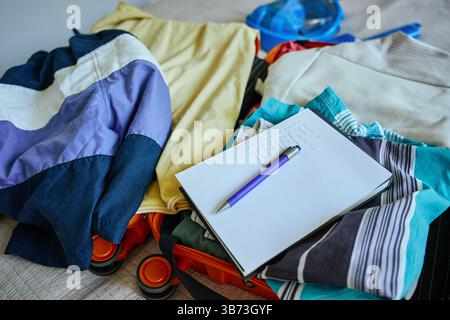 Elenco di controllo scritto a mano e penna sopra i vestiti piegati colorati all'interno della valigia aperta. Concetto di pianificazione del viaggio e di tutto il necessario per il viaggio Foto Stock