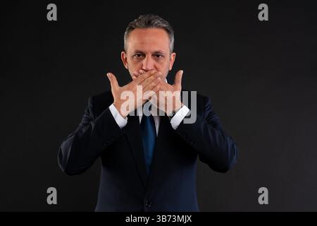 Un uomo d'affari maturo in tuta copre la bocca con le mani, esprimendo silenzio, segretezza o riservatezza. Foto Stock