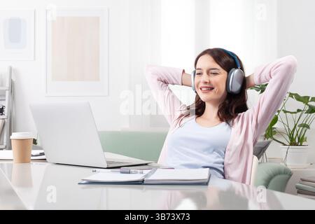 Giovane donna rilassata e sorridente a casa, seduta a una scrivania con le cuffie e navigazione sul suo notebook. Concetto di lavoro remoto positivo, studio online, vid Foto Stock