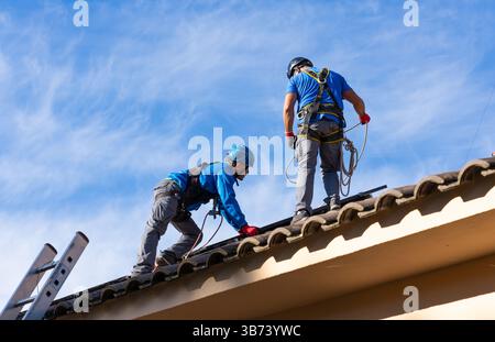 Due tecnici che lavorano sul tetto indossando attrezzature di sicurezza stanno installando pannelli fotovoltaici in un edificio residenziale Foto Stock
