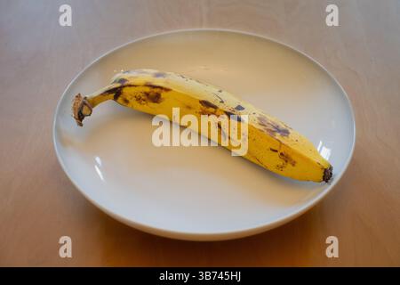 Una banana matura in un piatto bianco su un tavolo di legno Foto Stock