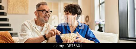 Una coppia di mezza età condivide risate e popcorn su un comodo divano di casa in un momento di gioia, striscione Foto Stock