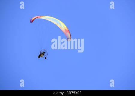 Paramotore con paracadute colorato che vola attraverso un cielo blu luminoso a Benotoa Beach Foto Stock