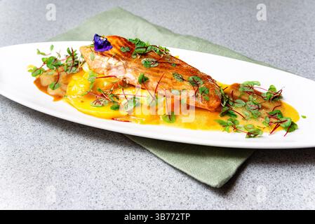 Filetto di salmone scottato servito su un letto di salsa di mango, guarnito con erbe fresche e fiori commestibili su un piatto bianco. Foto Stock