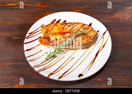 Un filetto di salmone perfettamente grigliato si trova su un piatto bianco, ricoperto di glassa balsamica e decorato con un rametto di rosmarino fresco, che presenta un delig Foto Stock