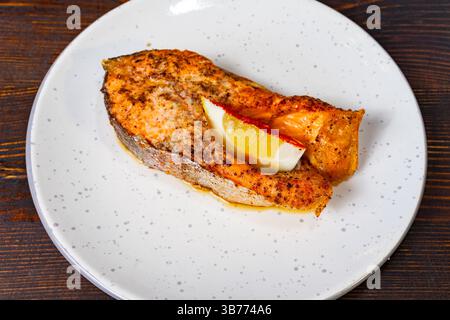 Filetto di salmone scottato in padella presentato su un piatto bianco guarnito con una fetta di limone. Il piatto presenta una crosta dorata, che indica un condimento saporito Foto Stock