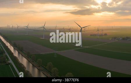 Una vista aerea panoramica delle turbine eoliche al tramonto, con campi verdi e corsi d'acqua che mostrano energia rinnovabile Foto Stock