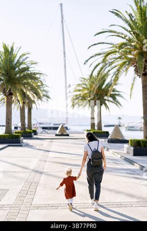 La donna cammina mano nella mano con la figlia di 2 anni tra le palme su un molo per barche. Foto Stock