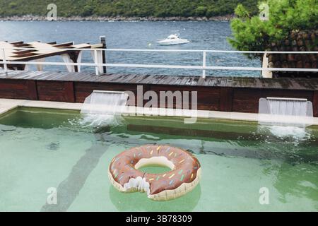 Un cerchio gonfiabile per nuotare galleggia in una piscina che si affaccia sul mare, sulle montagne e su uno yacht ormeggiato. Foto Stock