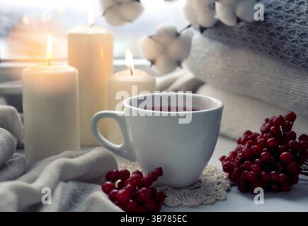 Tazza da tè con frutti di bosco decorata con maglie e candele su tavolo in legno, stile di vita sano, accogliente bevanda scandinava serale. Bevanda calda nella tazza. Foto Stock