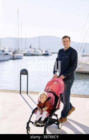 L'uomo sta spingendo sua figlia in un passeggino rosa su un molo. Foto Stock