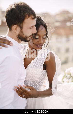 Coppia di nozze interrazziale. Matrimonio a Firenze, Italia. Lo sposo caucasico e la sposa afroamericana si coccolano sul tetto, al sole Foto Stock
