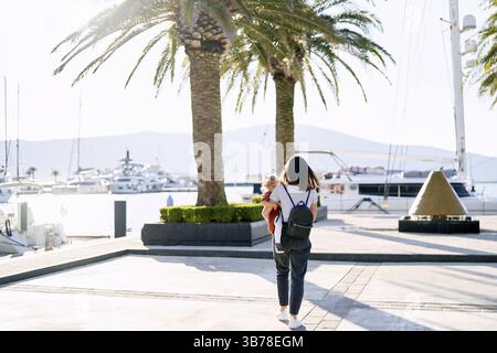 Una donna con una maglietta a righe e un jeans sta portando sua figlia tra le sue braccia tra le palme in riva al mare. Foto Stock