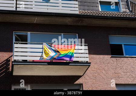Una vibrante bandiera d'orgoglio pende da un balcone in un'area residenziale, che mostra colori arcobaleno e simboli aggiuntivi. Il sole proietta un caldo bagliore Foto Stock
