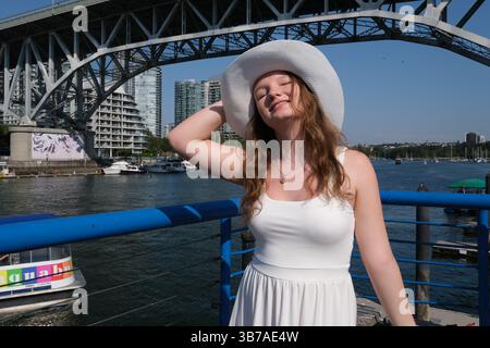 Donna eco-attivista hippie, viaggiatrice siede su un ponte vicino a un lago con le braccia allungate con un cappello e sorride sinceramente. Foto di alta qualità Foto Stock