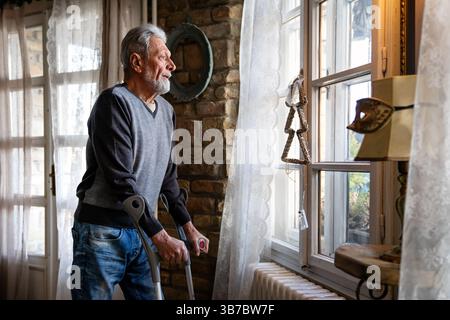 Vecchio con la stampella che si sente deluso, perso in pensieri tristi da solo a casa. Vedova uomo maturo. Foto Stock