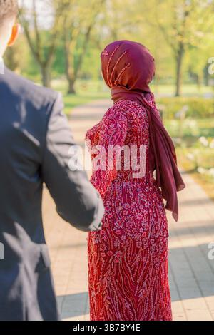 Una coppia cammina attraverso un parco illuminato dal sole, tenendosi per mano con un gesto intimo. La donna indossa un abito rosso dettagliato e un velo, mentre l'uomo lo è Foto Stock