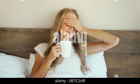 Donna stanca che si sveglia dopo aver dormito lunedì tenendo una tazza di caffè, toccando la testa, soffrendo di mal di testa, stress, affaticamento, emicrania adagiata Foto Stock