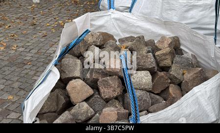 Grandi borse bianche da costruzione piene di ciottoli grigi su strada acciottolata con foglie autunnali cadute in un ambiente urbano. Foto Stock