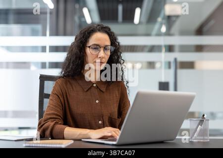 Donna professionale concentrata con capelli ricci che lavora su un notebook in un ufficio moderno. Sta indossando gli occhiali. Foto Stock