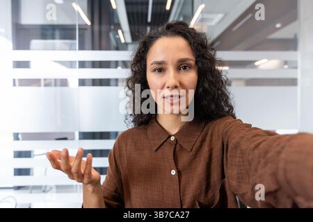 Una donna con i capelli ricci mentre parla in una fotocamera in un ufficio moderno. Lo sfondo presenta pannelli di vetro e luci luminose. Foto Stock