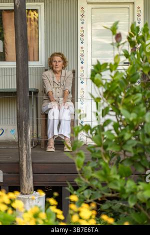 Donna anziana seduta comodamente su una sedia sul portico della sua casa di campagna, assaporando la tranquillità e godendo la bellezza dei fiori e della lussureggiante gr Foto Stock