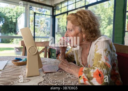 Donna anziana seduta a un tavolo nella sua accogliente casa, applicando il rossetto rosso con un piccolo specchio, abbracciando la sua routine di bellezza e preparandosi con sicurezza Foto Stock