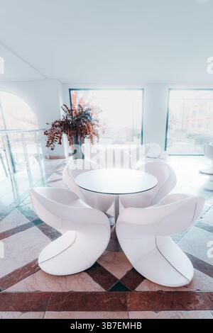 Interni moderni e minimalisti con futuristiche sedie bianche e un tavolo rotondo in un'elegante area lounge, con pavimenti in marmo, grandi finestre e decorazioni Foto Stock