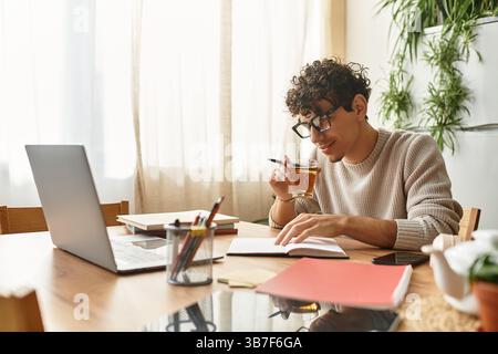 Un bell'uomo prende un sorso premuroso dalla sua tazza mentre scrive nel suo taccuino a casa. Foto Stock