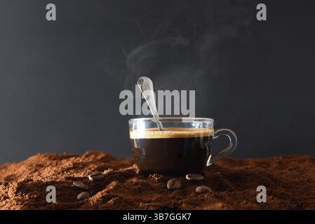 Dettaglio di una tazza di caffè in vetro con caffè caldo appena estratto e cucchiaio di metallo all'interno su una pila di caffè macinato con chicchi intorno e iso scuro Foto Stock