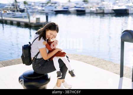 La giovane donna sta abbracciando sua figlia stretta mentre si siede su un molo in un porticciolo. Foto Stock