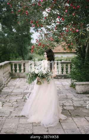 La sposa si trova sotto un oleandro in fiore con un bouquet di rose bianche, peonie, delfini e rami di eucalipto nelle sue mani. Foto Stock