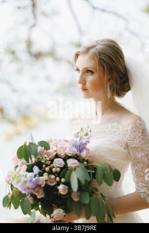 Una sposa sofisticata si erge con un bouquet nuziale nelle sue mani sotto i rami di un albero, primo piano. Foto Stock