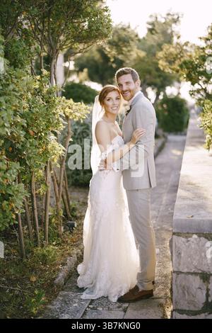 Lo sposo abbraccia la sposa in piedi sul vicolo vicino ai cespugli verdi. Foto Stock