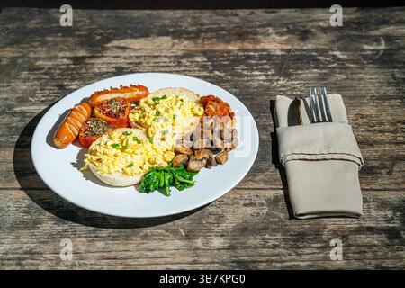 Colazione abbondante con pane, uova strapazzate, salsicce, pomodoro arrosto, fagioli al forno, funghi saltati e spinaci su piatto bianco. Isolato da un tavolo di legno Foto Stock
