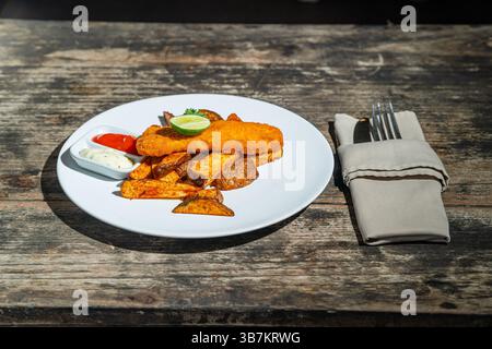 Pesce fritto e patatine fritte, servite con remoulade (salsa, senape, erbe aromatiche) su piatto bianco. Isolato da un tavolo di legno. Fotografia gastronomica Foto Stock