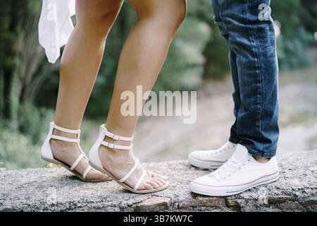 I piedi femminili in sandali accanto ai piedi maschili in sneakers si stagliano su un bordo di pietra nel parco. Foto Stock