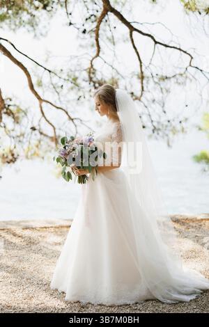 Una sposa sofisticata si erge con un bouquet di nozze nelle sue mani sotto i rami di un albero sul mare . Foto Stock