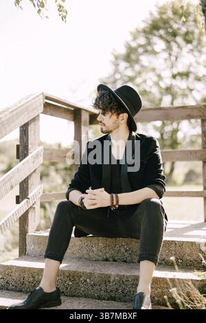 Un giovane con cappello nero siede su gradini di pietra con ringhiere in legno. Foto Stock
