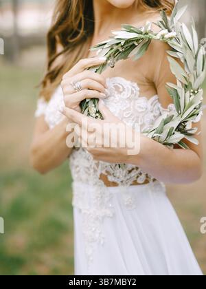 La splendida sposa con un abito bianco ricamato si stende lateralmente e tiene in mano una corona di rami verdi di ulivo. Foto Stock