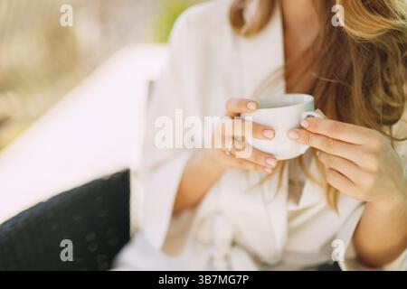 Una donna in vestaglia al mattino siede su una sedia e tiene una tazza in mano . Foto Stock