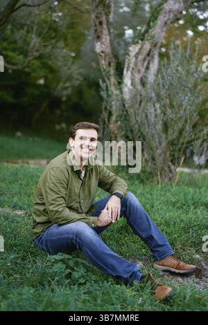 Un giovane sorridente seduto sull'erba verde nel parco. Foto Stock