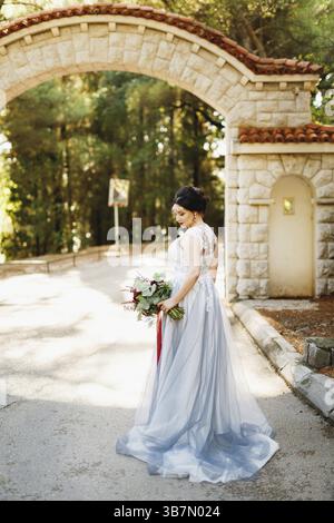 Una sposa con un bouquet nuziale si erge sotto un arco di mattoni all'ingresso del parco. Foto Stock